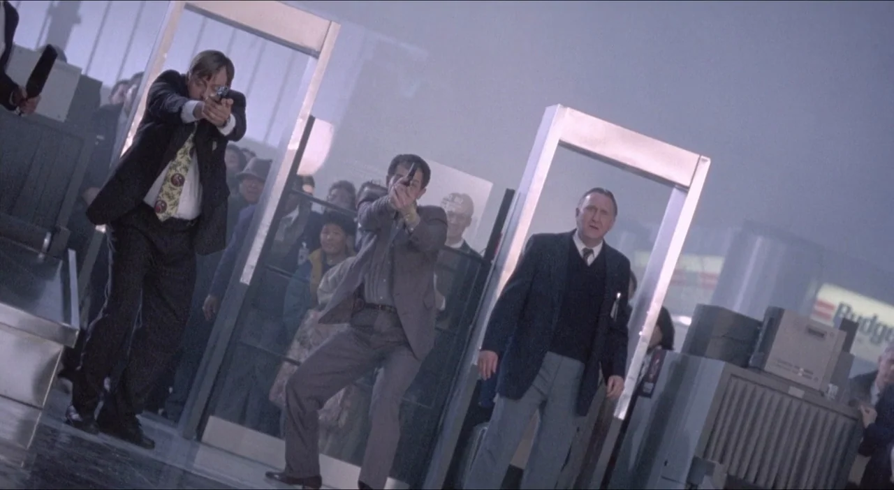 Twelve Monkeys (1995) movie still: fullbody — A busy airport terminal security checkpoint features metal detectors and X-ray conveyor belts. Two men…