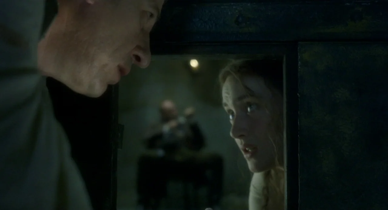 Quills (2000) movie still: medium — A dimly lit room with a dark frame separating two individuals in shadow. A woman gazes through the rectangular…