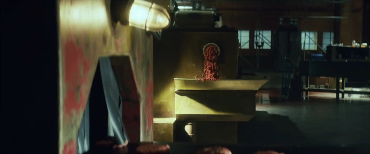 American Carnage (2022) movie still: medium — An industrial food processing machine sits in a dimly lit factory room. Raw ground meat is actively…
