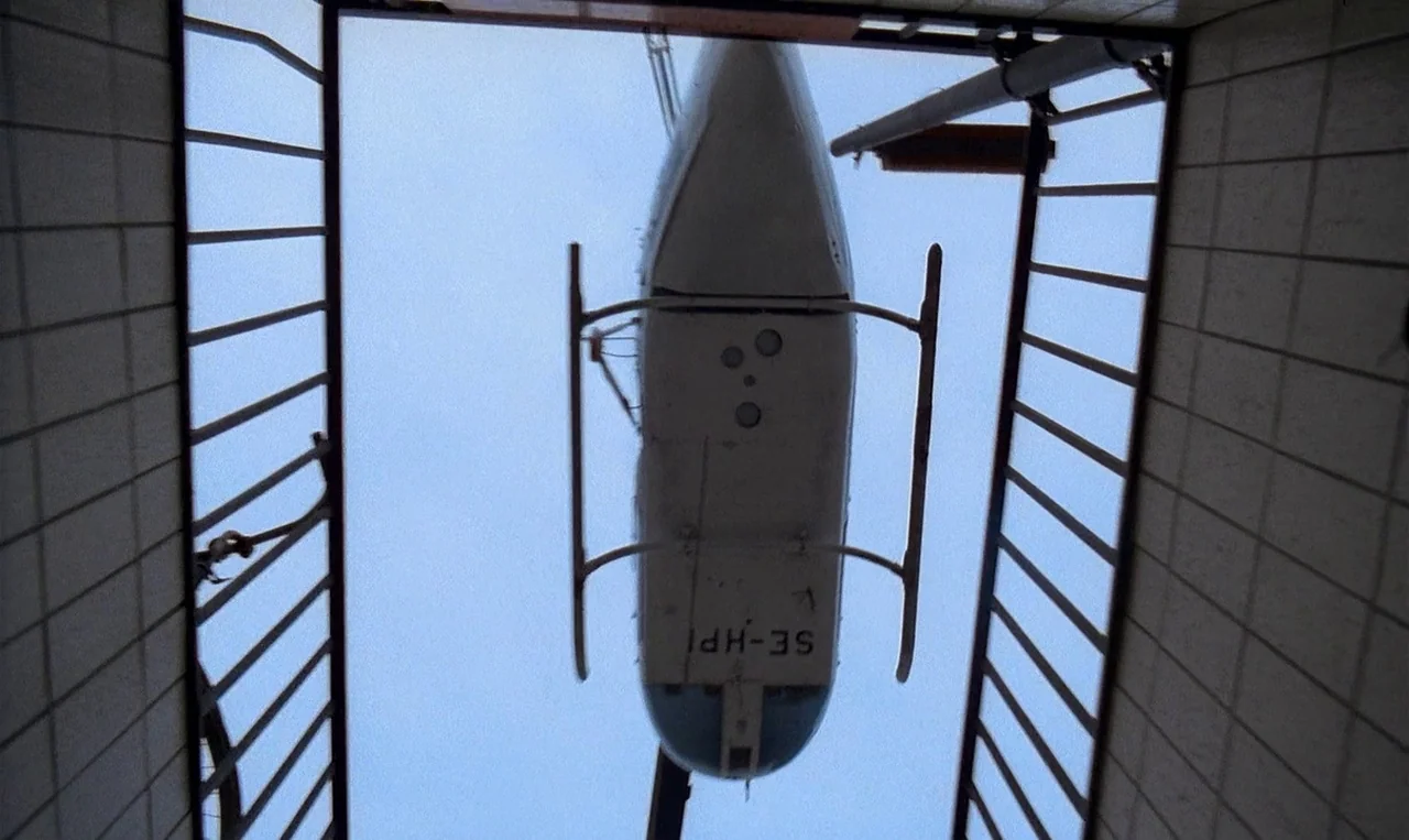Man on the Roof (1976) movie still: wide — A low-angle view looks through a tiled stairwell opening toward the sky. A white light helicopter is…