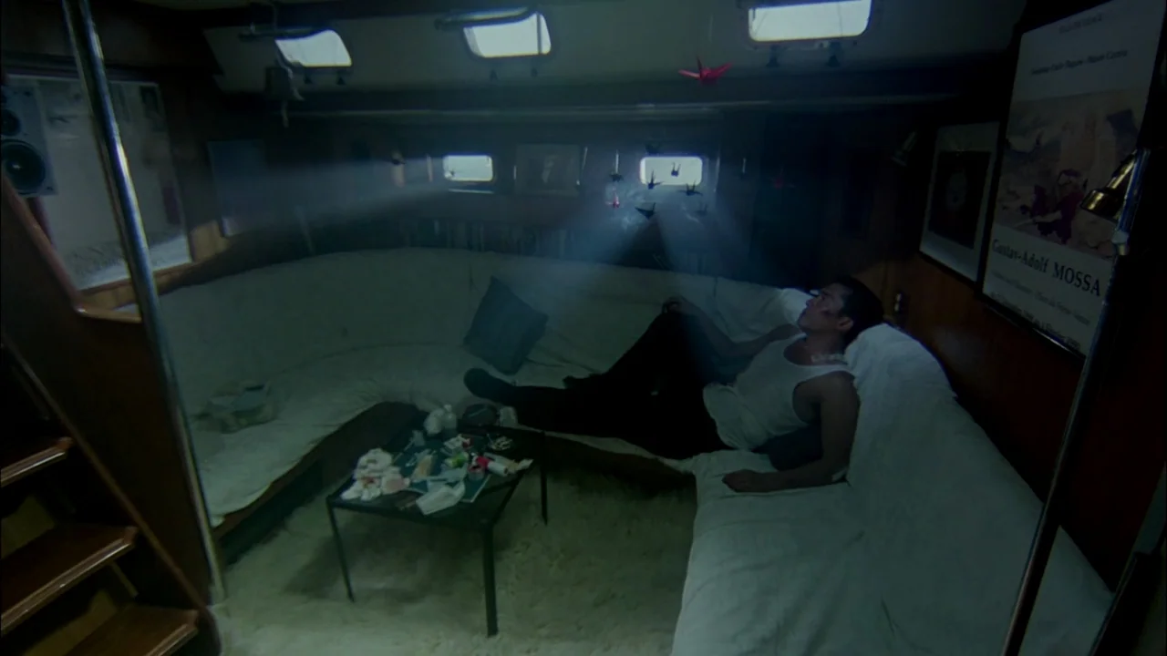 Hard Boiled (1992) movie still: wide — A dimly lit, cramped cabin on a sailboat features white cushioned seating and small windows. A man reclines…