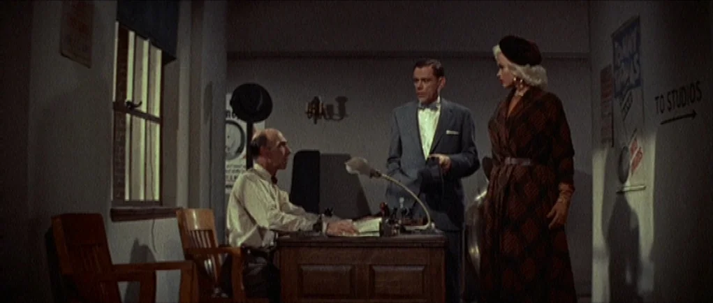 The Girl Can’t Help It (1956) movie still: fullbody — A dimly lit room functions as an administrative space with wood furniture and signage. Three…