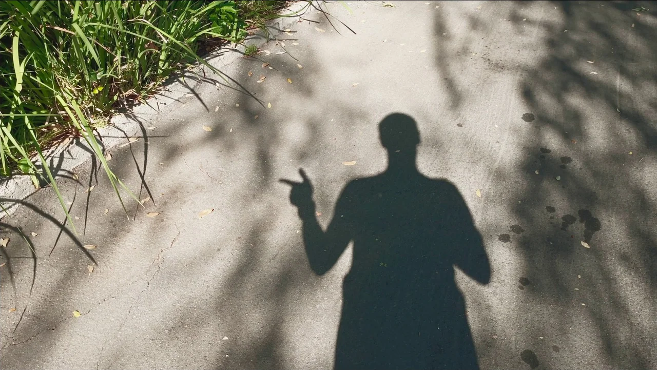 Transformers: Rise of the Beasts (2023) movie still: wide — An asphalt path is bordered by tall green grass and scattered dry leaves. A dark human…