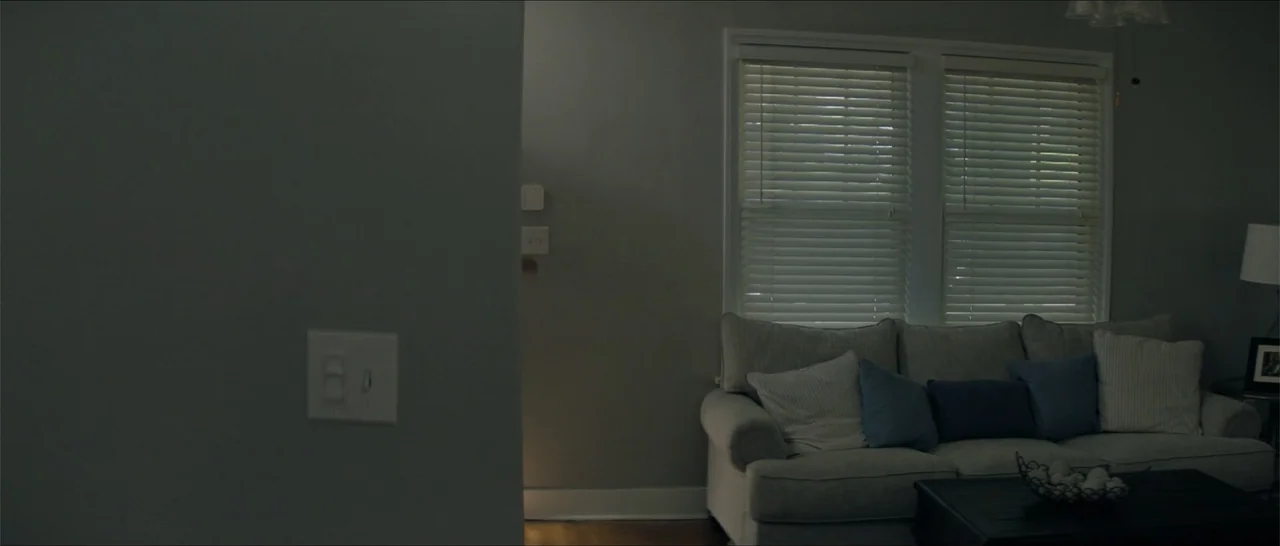 Crescent City (2024) movie still: wide — A modern living room features a grey couch with blue pillows and closed blinds. A dark coffee table sits in…