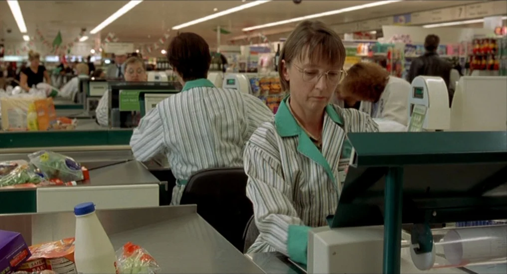 All or Nothing (2002) movie still: medium — A busy checkout area in a grocery store with rows of registers and fluorescent lights. A cashier wearing…
