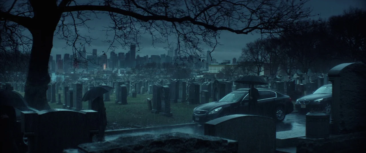 John Wick (2014) movie still: wide — A rain-slicked cemetery filled with stone markers sits before a distant city skyline under clouds. Two figures…