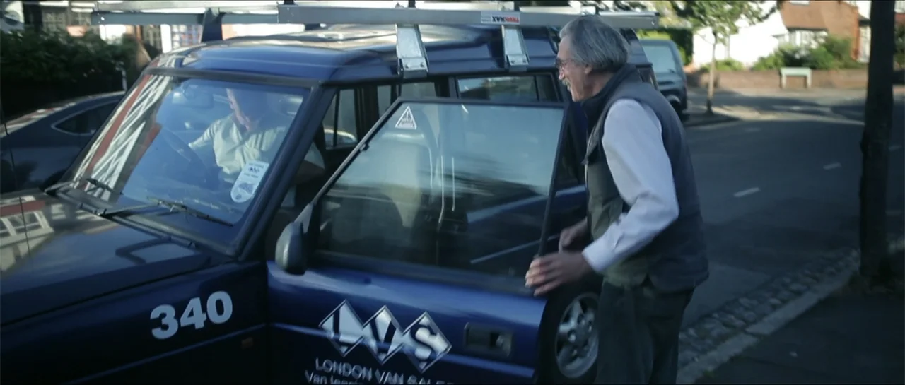 Garnet’s Gold (2014) movie still: medium — A dark blue van with ladders on the roof is parked on a suburban road. An older man in a grey vest stands…