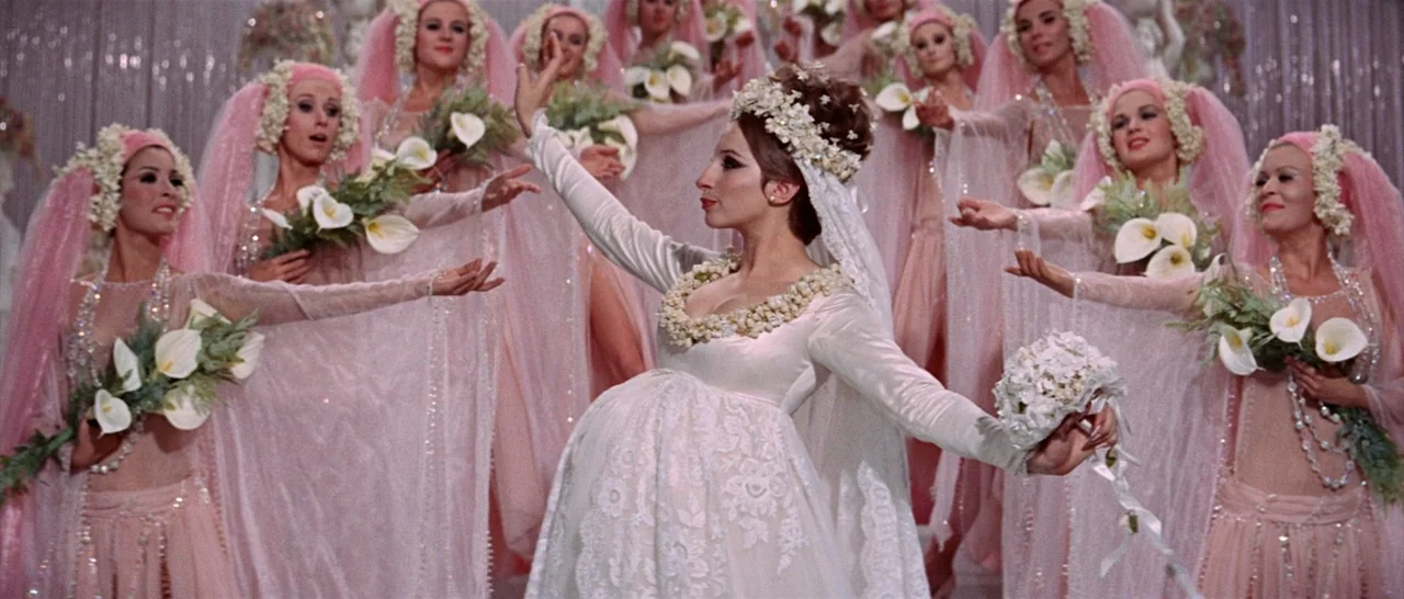 Funny Girl (1968) movie still: medium — A theatrical stage set decorated with white curtains and hanging floral arrangements. A woman in a wedding…