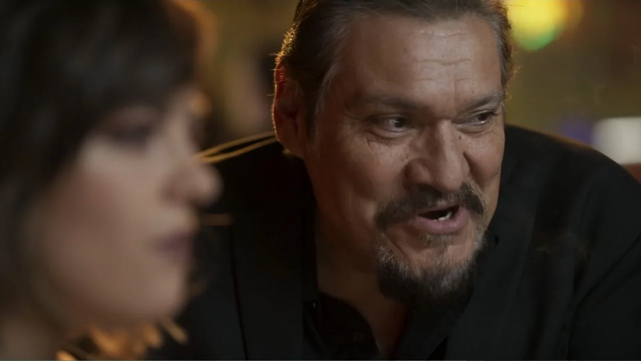 Lecciones para canallas (2022) movie still: closeup — A dimly lit bar interior features a man in a black shirt speaking intensely. A woman is…