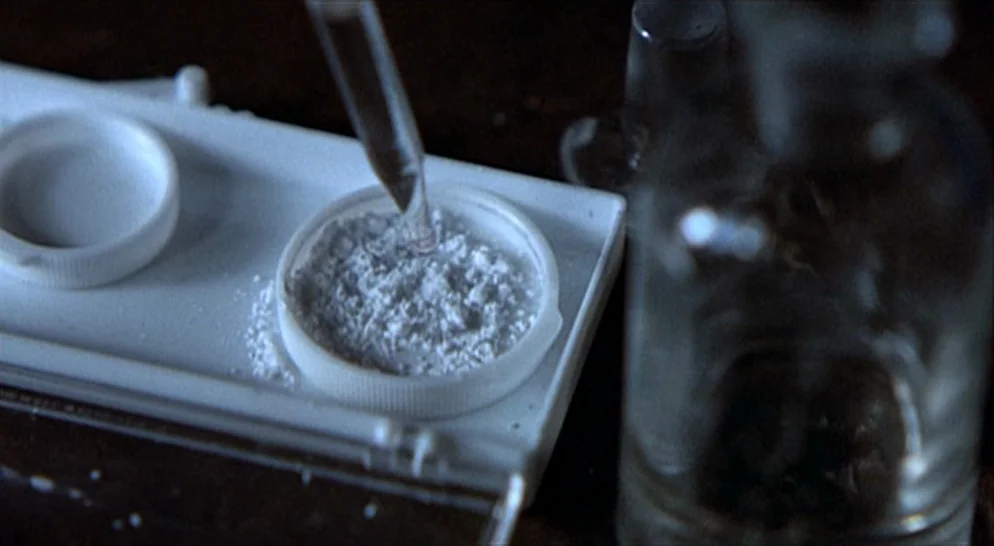 The French Connection (1971) movie still: closeup — A white plastic tray sits on a dark surface containing a pile of white powder. A glass pipette…