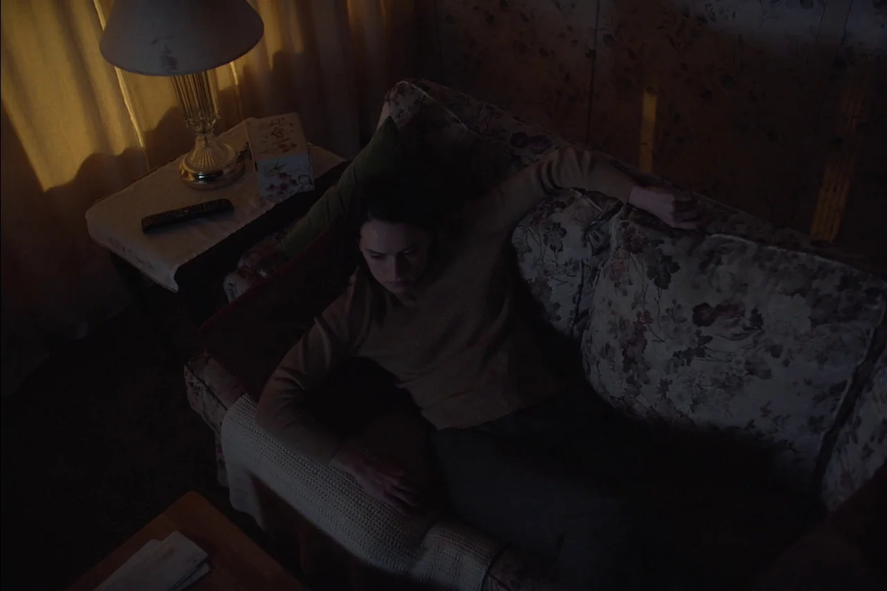 Sometimes I Think About Dying (2023) movie still: medium — A dimly lit room with floral-patterned upholstery and a single warm table lamp. A woman…