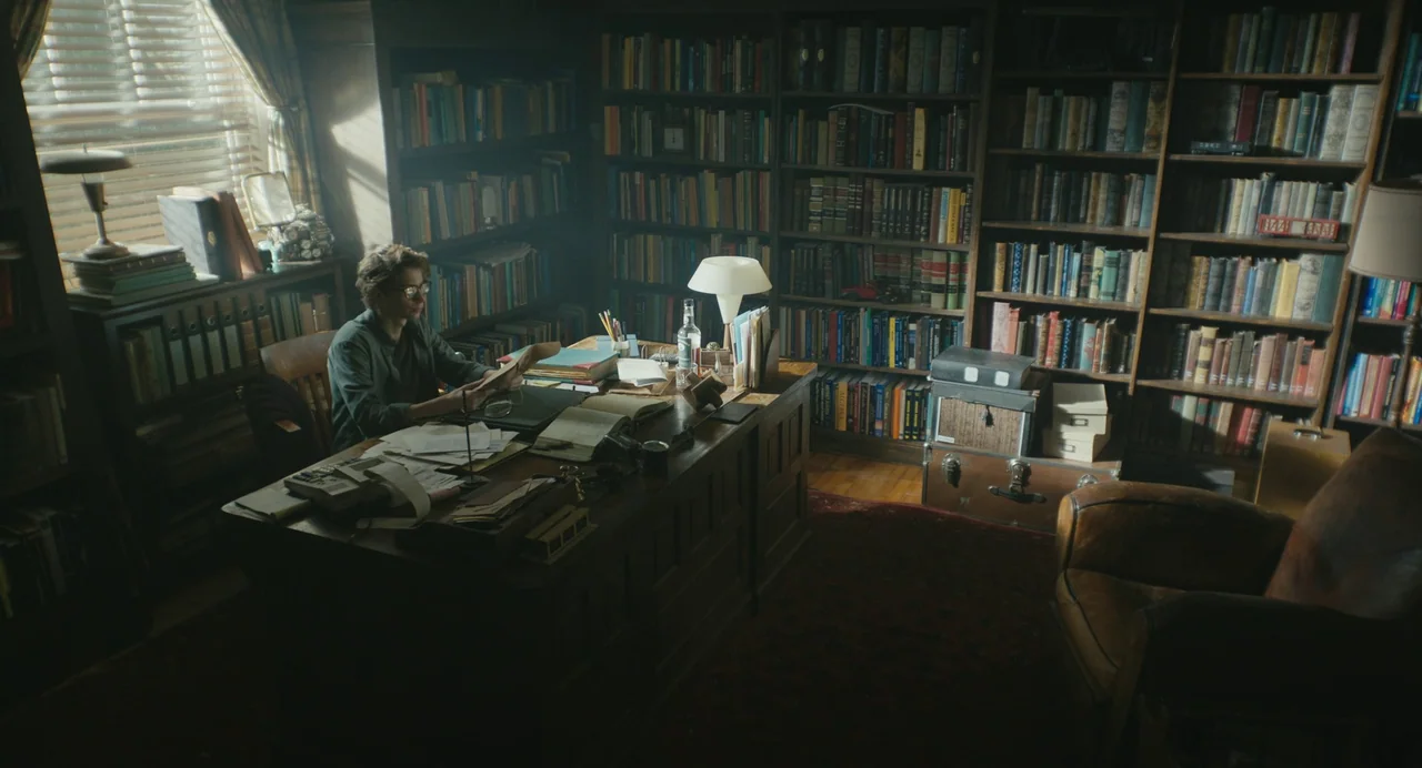 The Life of Chuck (2024) movie still: wide — A room filled with wall-to-wall bookshelves and a large wooden desk holding papers. A man sits at the…