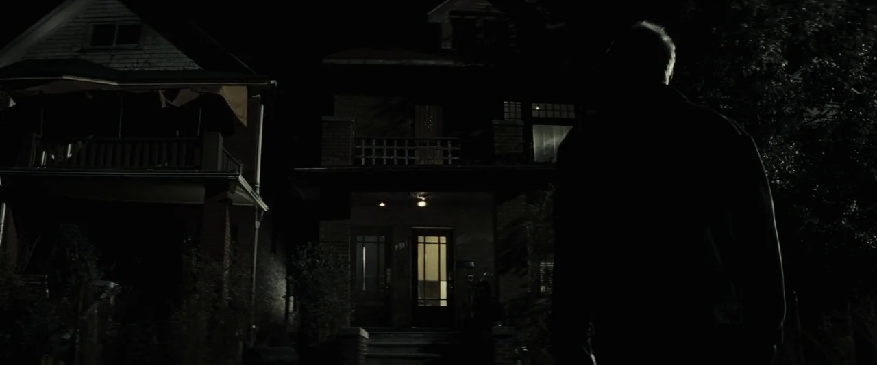 Gran Torino (2008) movie still: medium — A residential street at night shows a silhouette of a man viewing dark houses. Porch lights illuminate the…