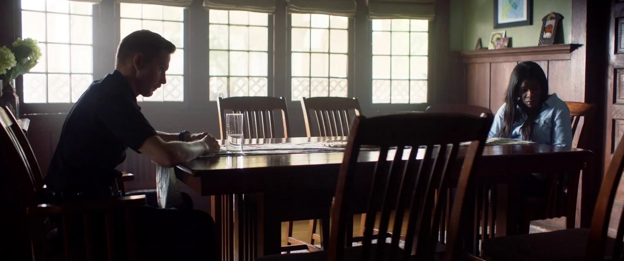 Blindfire (2020) movie still: medium — A dark wood dining room features a long table centered between large grid windows. A man and a woman sit at…