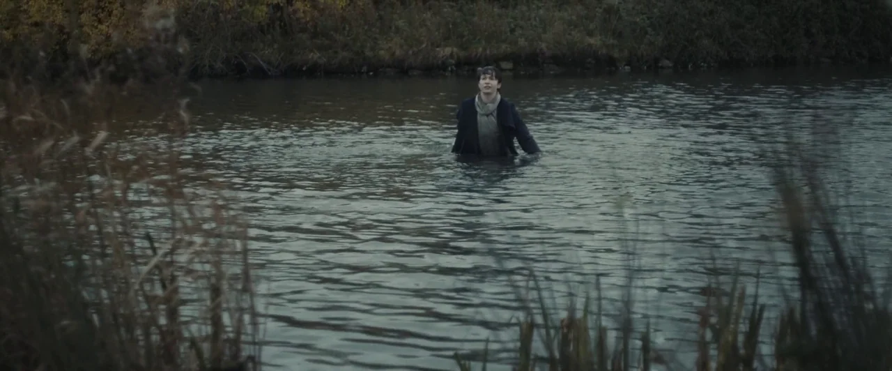 You Are Not My Mother (2021) movie still: wide — A woman stands in the middle of a river with dense brush on the banks. The water is murky and dark…