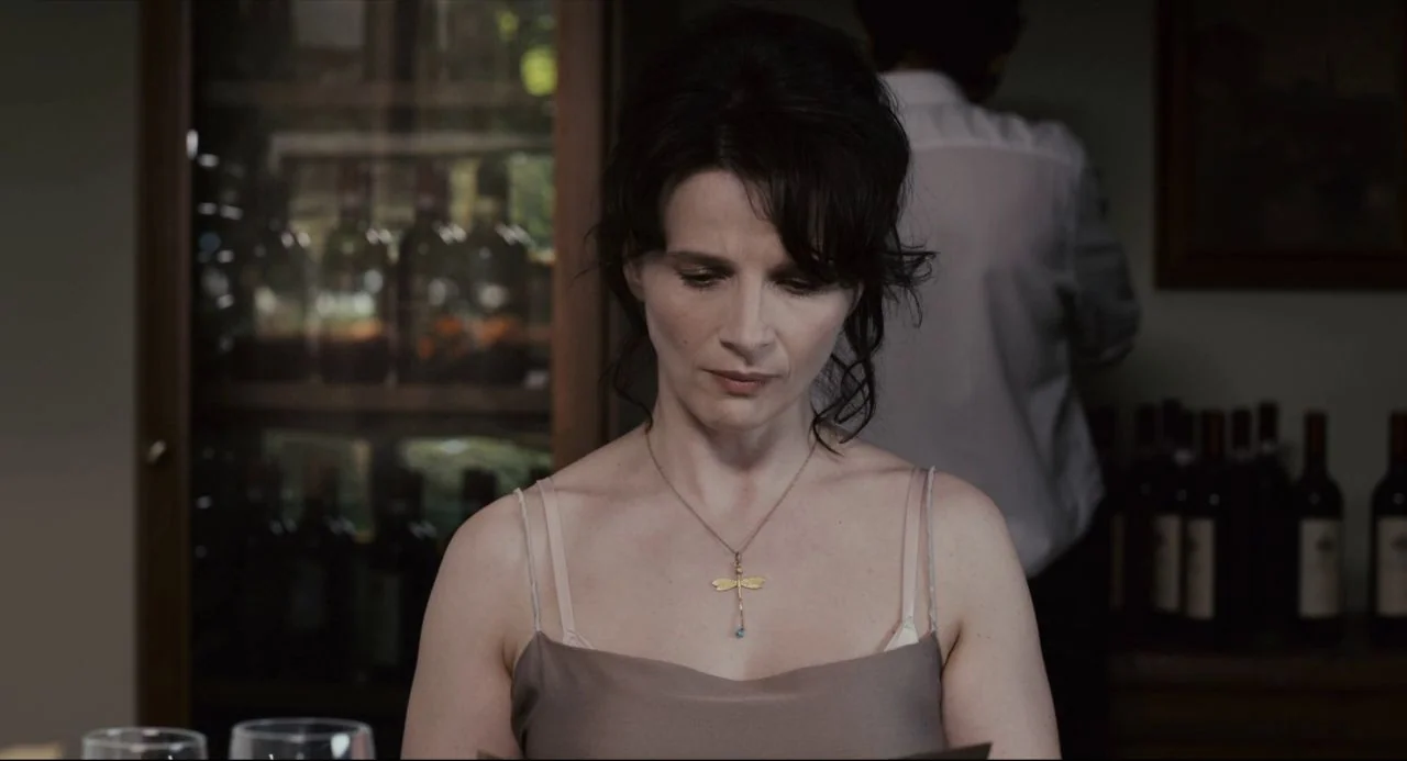 Certified Copy (2010) movie still: medium — A dimly lit indoor dining area contains a glass-fronted wine cabinet in the background. A woman in a tan…