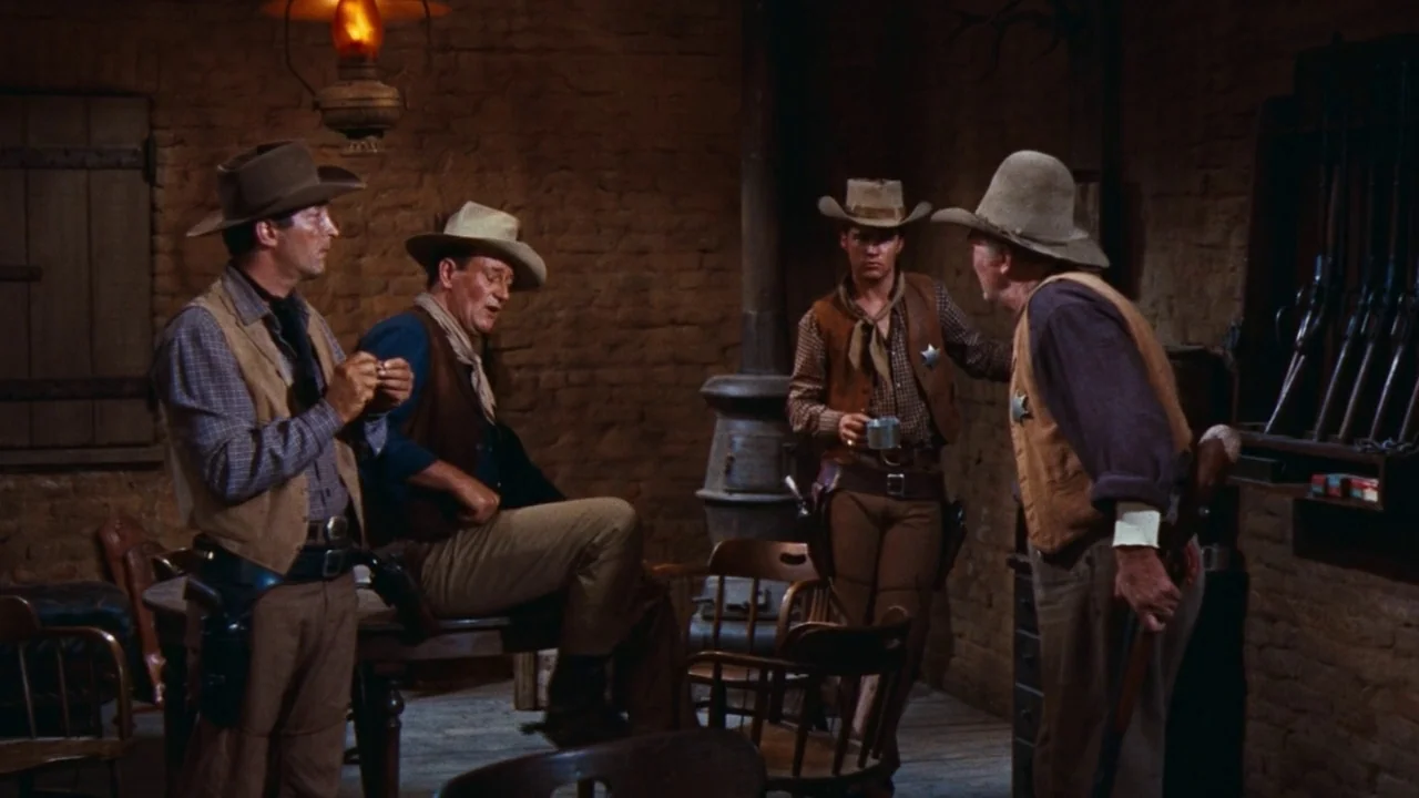 Rio Bravo (1959) movie still: medium — A rustic office with textured mud-brick walls contains wooden furniture and a stove. Four men in mid-century…