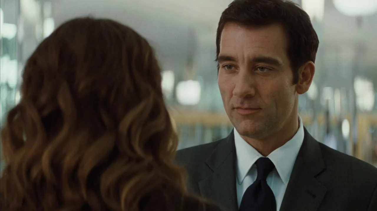 Duplicity (2009) movie still: medium — A modern office interior with blurred architectural features in the background. A man wearing a suit stands…