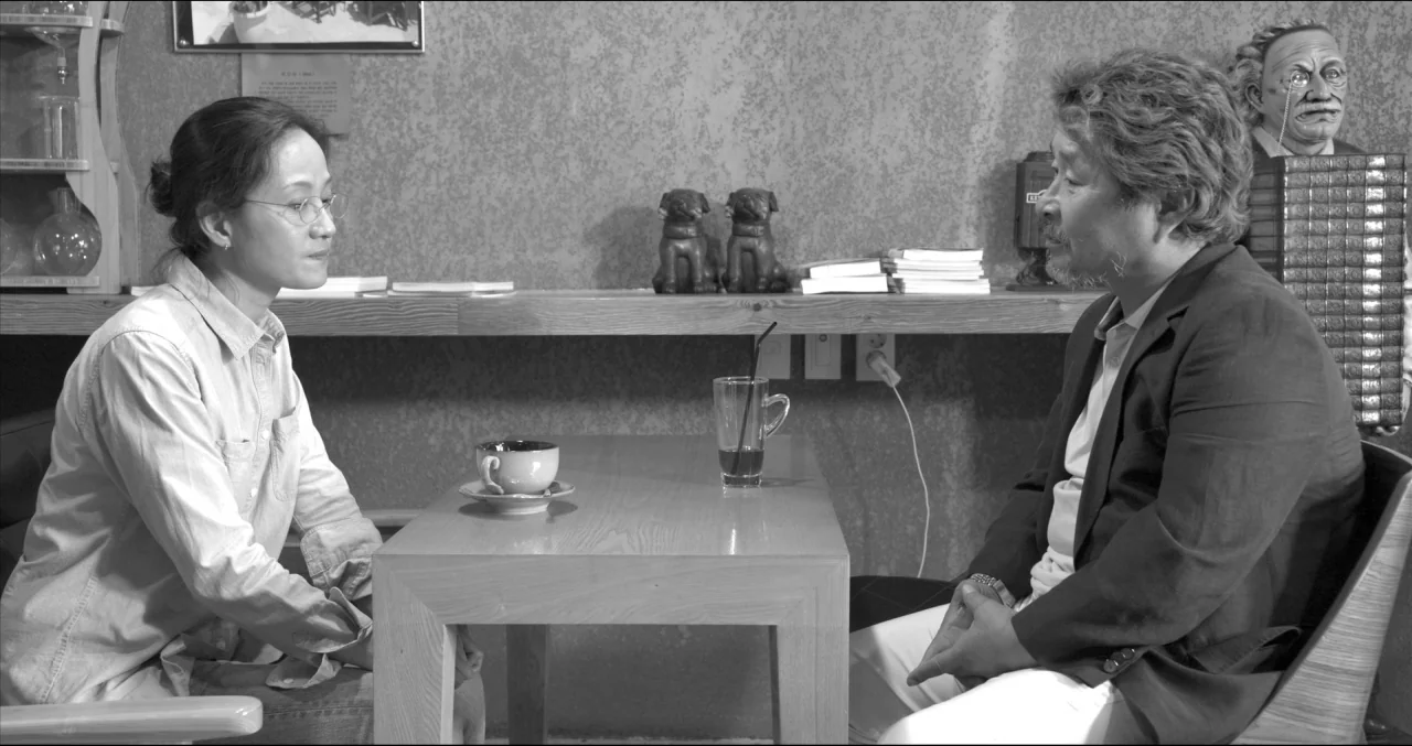 Grass (2018) movie still: medium — A man and a woman sit facing each other at a table in a dimly-lit cafe. A coffee cup, a glass with a straw, and a…