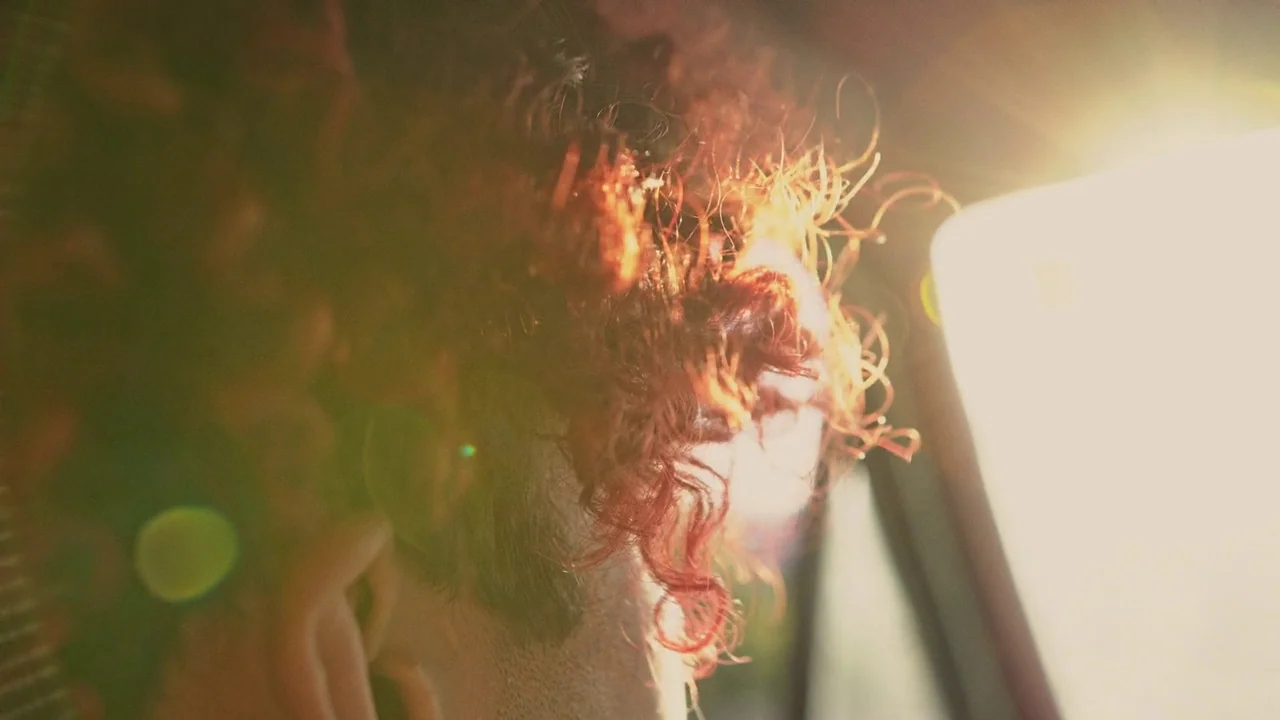 Gasoline Rainbow (2023) movie still: closeup — A close-up view shows the back of a red-haired person inside a vehicle cabin. Sunlight beams through…
