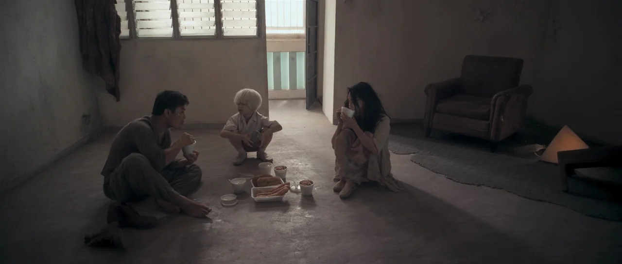 Rigor Mortis (2013) movie still: wide — A sparsely furnished room with concrete floors, bare walls, and boarded-up windows. A man, a young boy, and…