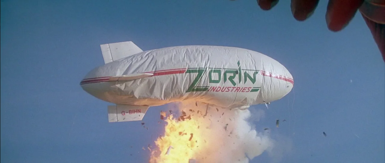 A View to a Kill (1985) movie still: wide — A large airship with branded text floats against a clear blue sky. The underside of the dirigible is…