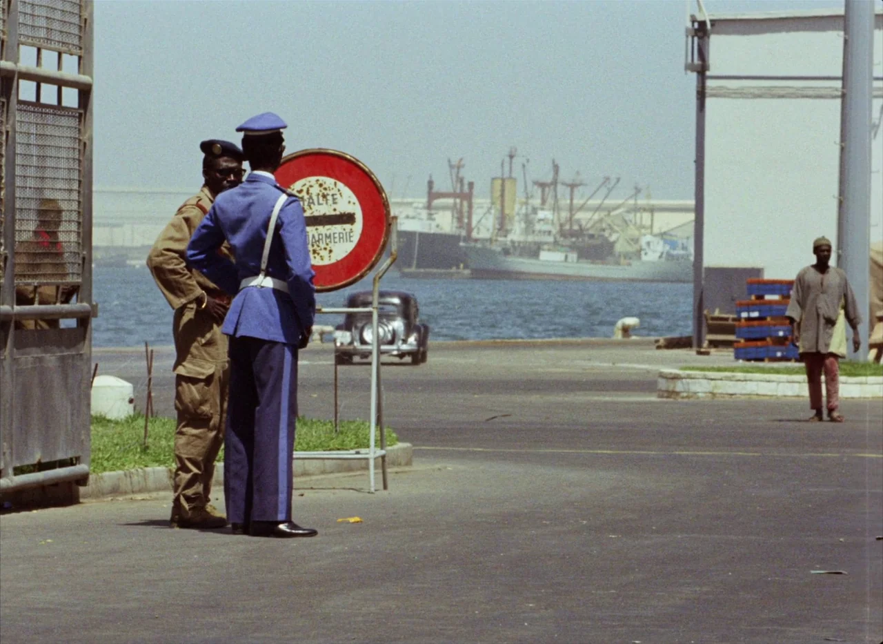 Touki Bouki (1973) movie still: fullbody — A bustling waterfront area with large cargo ships anchored in the distance. Two uniformed men stand by a…