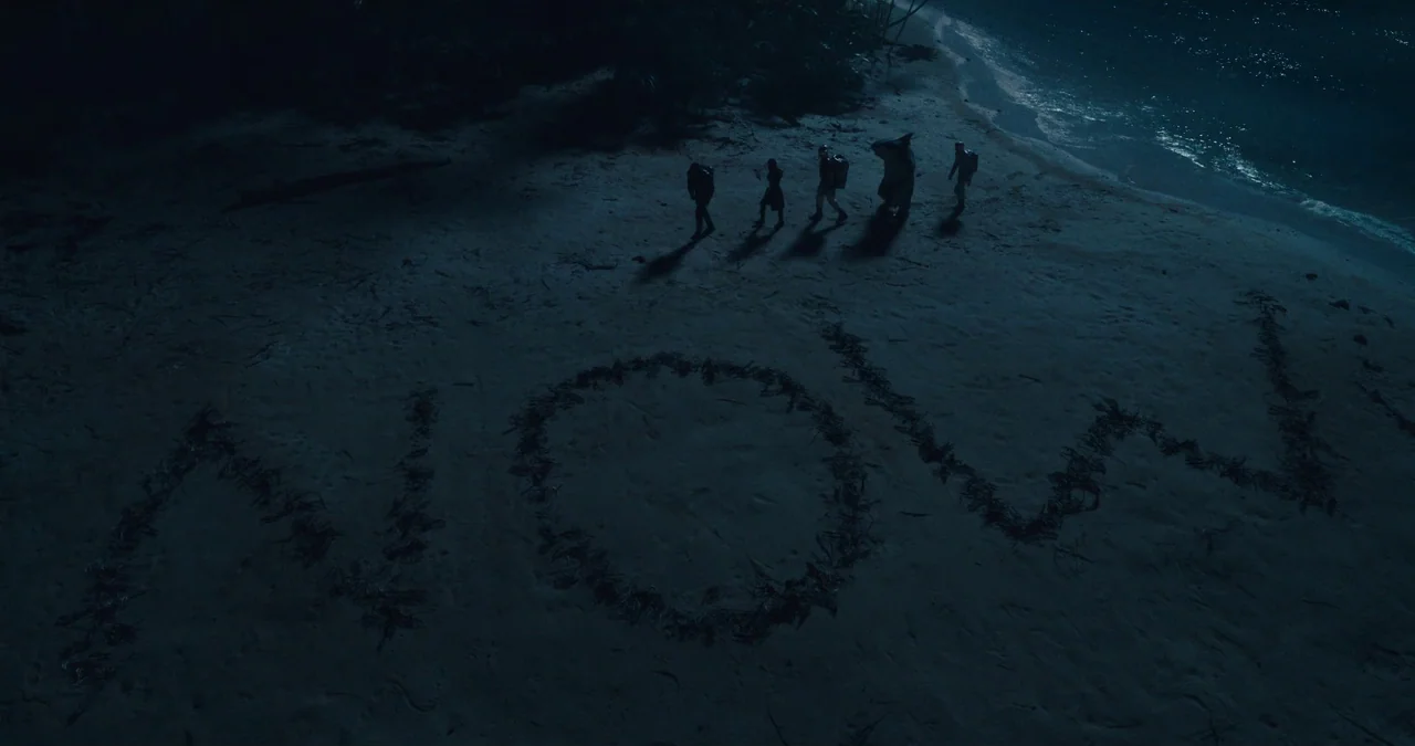 The Suicide Squad (2021) movie still: establishing — A dark tropical beach features the word 'NOW' written in large sand-etched letters. Five…