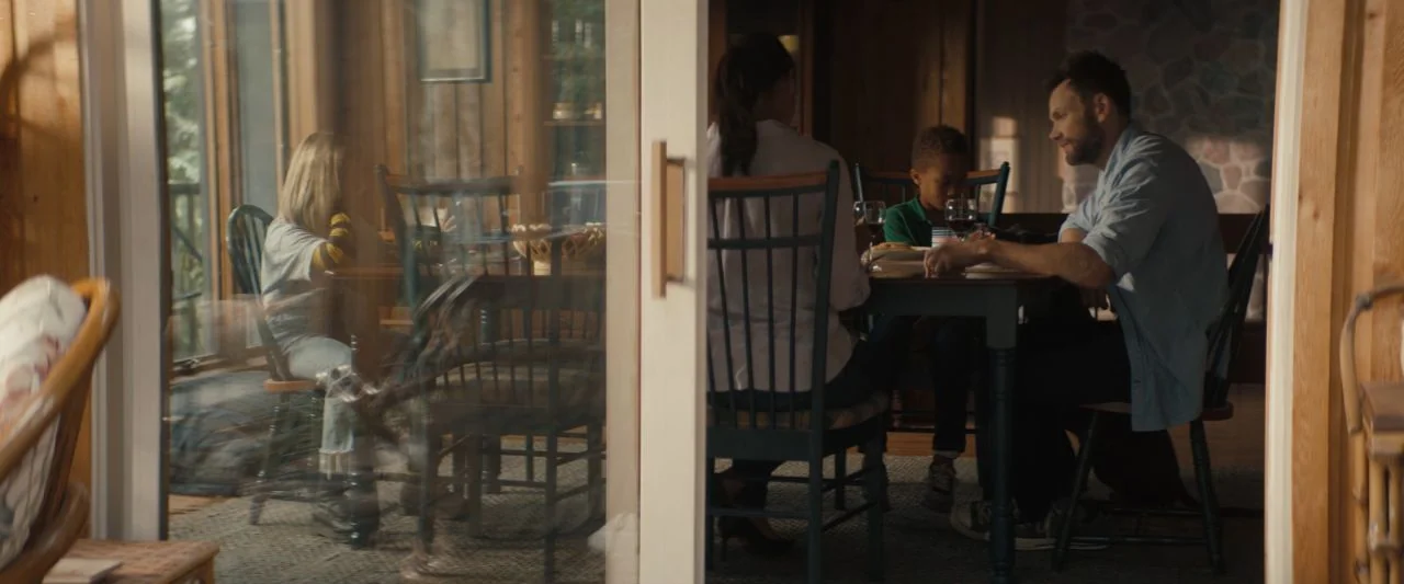 Becky (2020) movie still: wide — A rustic dining room features warm wood paneled walls and large floor-to-ceiling windows. A man, woman, and child…