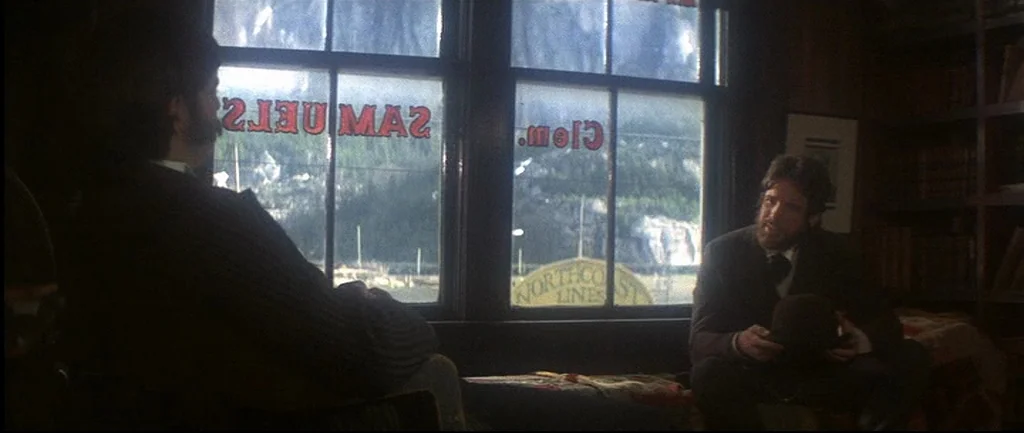 McCabe & Mrs.Miller (1971) movie still: medium — A dimly lit room with wooden walls overlooks a mountainous exterior through two windows. Two men in…