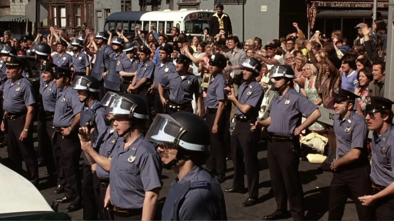 Dog Day Afternoon (1975) movie still: wide — A city street filled with a large crowd of people and police officers. A line of police officers…