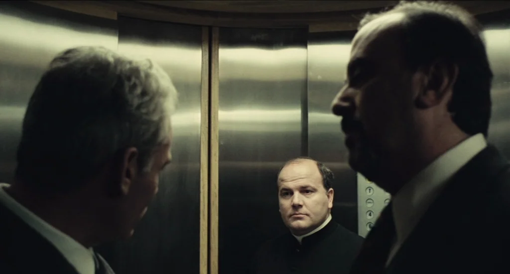 Todo Modo (1976) movie still: medium — Three men stand in a small, metallic elevator with brushed steel wall panels. A priest stands centrally in…