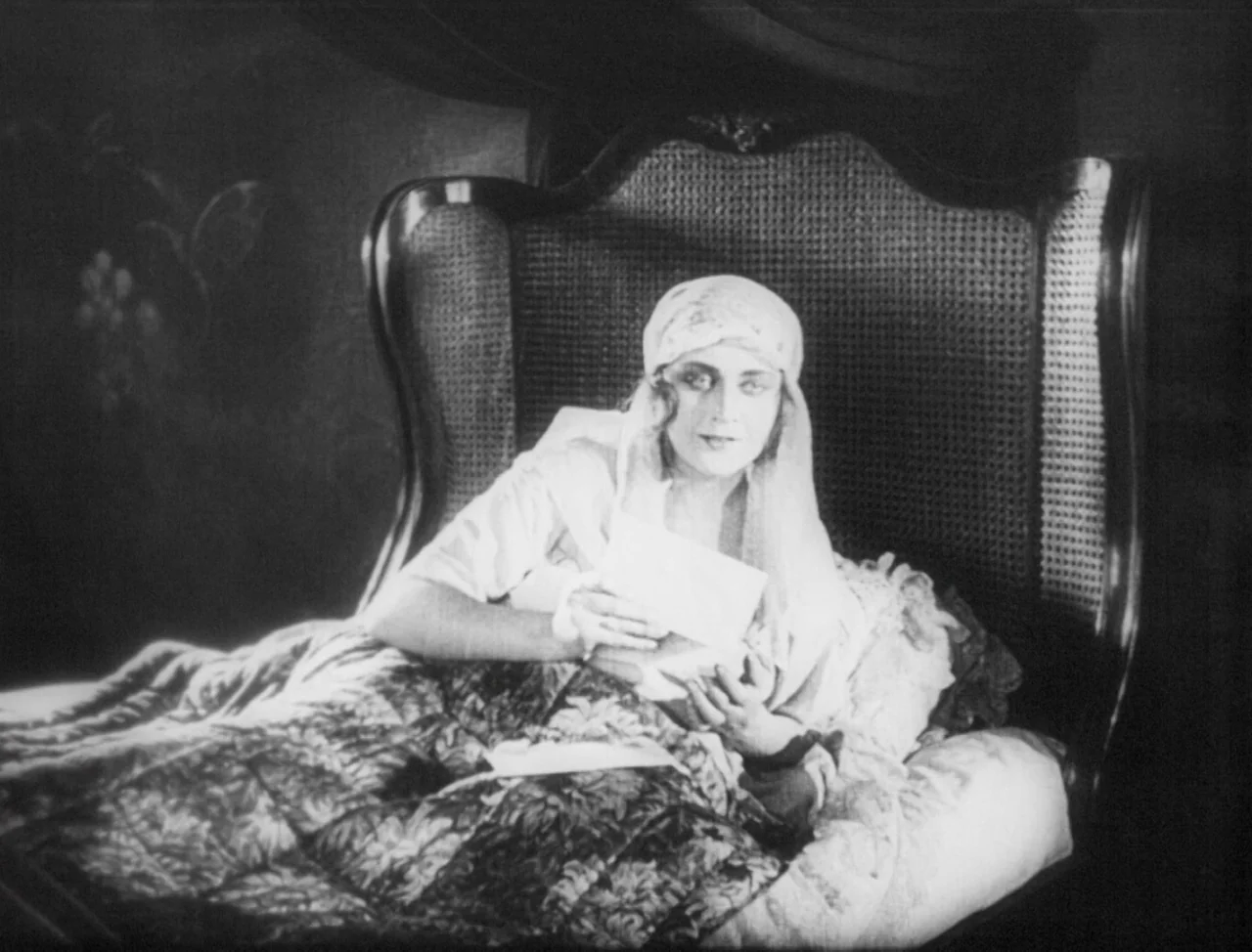 The Hands of Orlac (1924) movie still: medium — A woman sits in a dark room leaning against a large ornate headboard. She holds a small letter in…