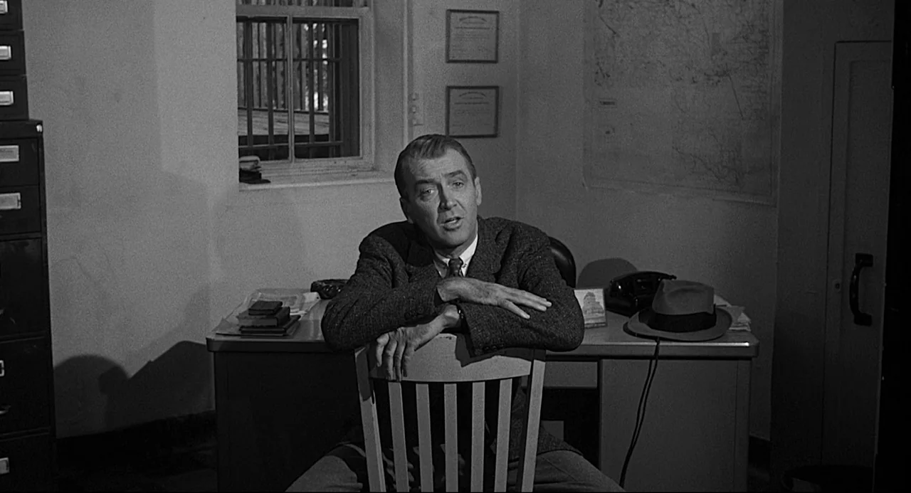 Anatomy of a Murder (1959) movie still: medium — An office features a metal desk, filing cabinets, and a barred window. A man in a suit sits…