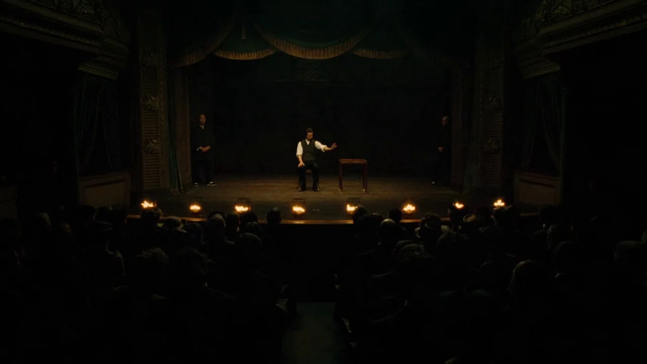 The Illusionist (2006) movie still: wide — A dimly lit old theater features a wooden stage with a performer and a table. A dark audience sits in the…