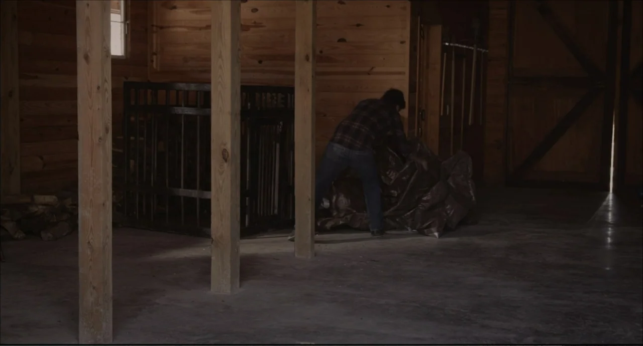 Freedom (2024) movie still: wide — An empty wooden barn structure features support pillars and a large metal cage. A man wearing a plaid shirt moves…