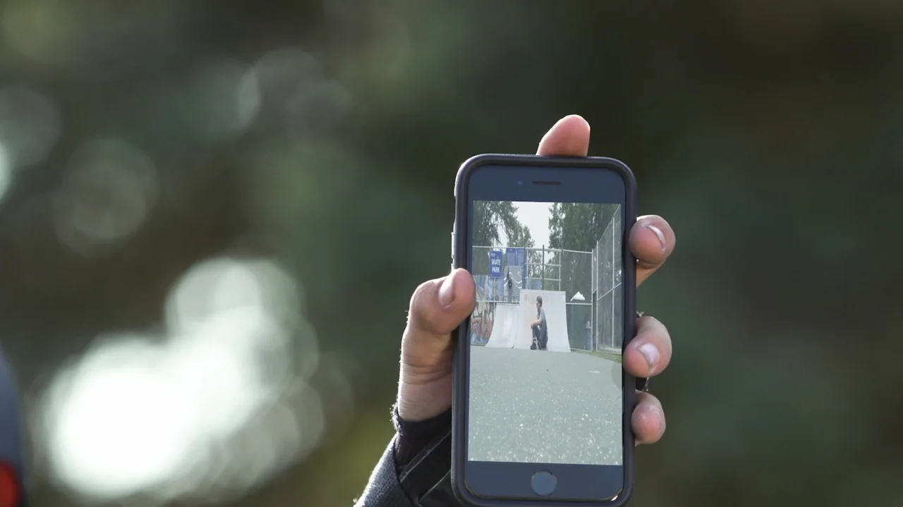 Sk8 Dawg (2018) movie still: closeup — A close-up view of a hand holding a smartphone in an outdoor setting. The screen displays a person sitting on…