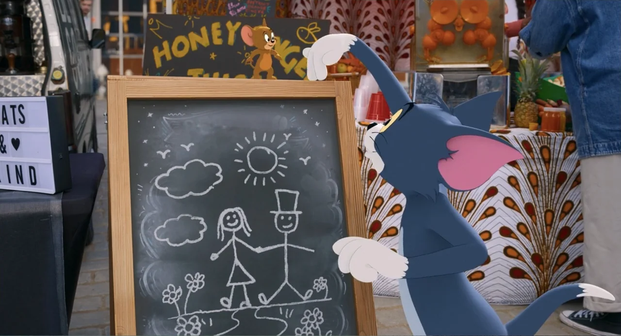 Tom & Jerry (2021) movie still: medium — An outdoor market stall features a blackboard with a stick figure drawing. A blue animated cat gestures at…