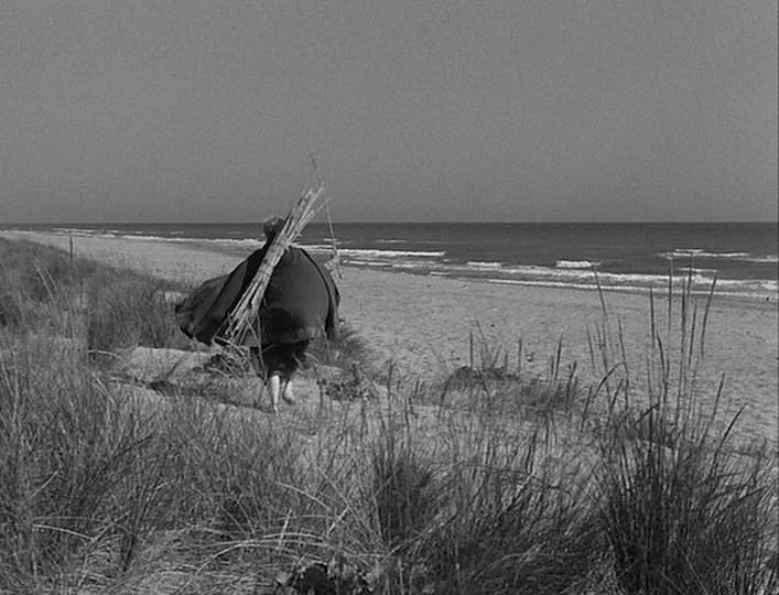 La Strada (1954) movie still: wide — A desolate sandy beach with tall beach grass in the foreground and the ocean. An elderly figure wearing a dark…