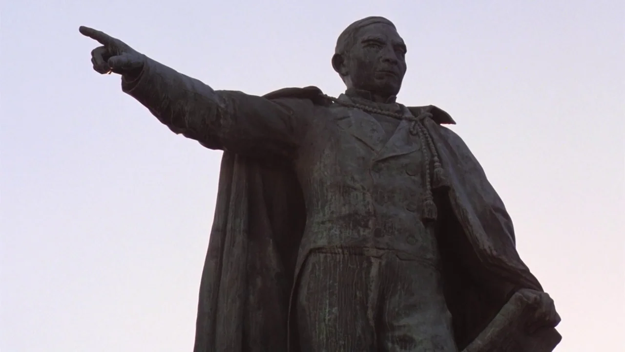 Being There (1979) movie still: medium — A bronze statue of a man stands against a bright, featureless pale sky. The figure is posed pointing a…