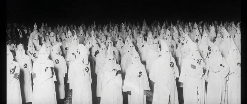 Manderlay (2005) movie still: wide — A large crowd of individuals stands in a dark outdoor setting at night. Everyone wears identical white robes…