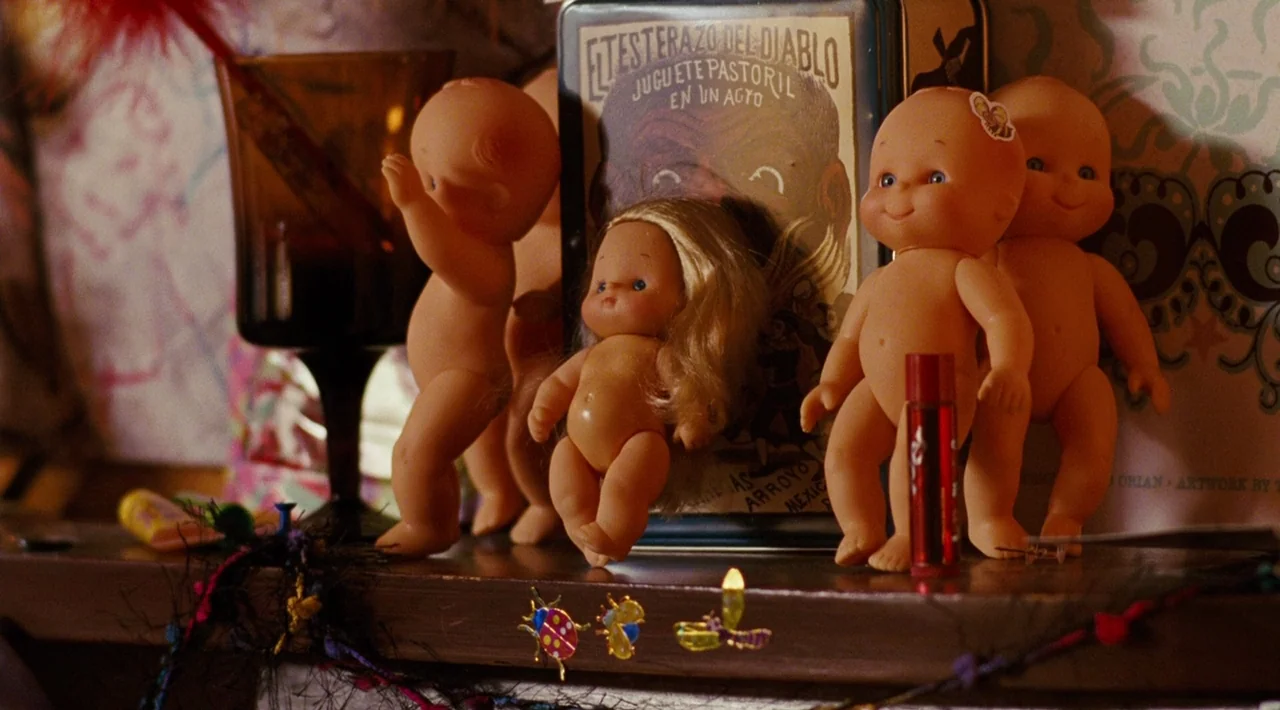 Juno (2007) movie still: closeup — A wooden shelf holds several small plastic baby dolls and a metal decorative tin. The background features…