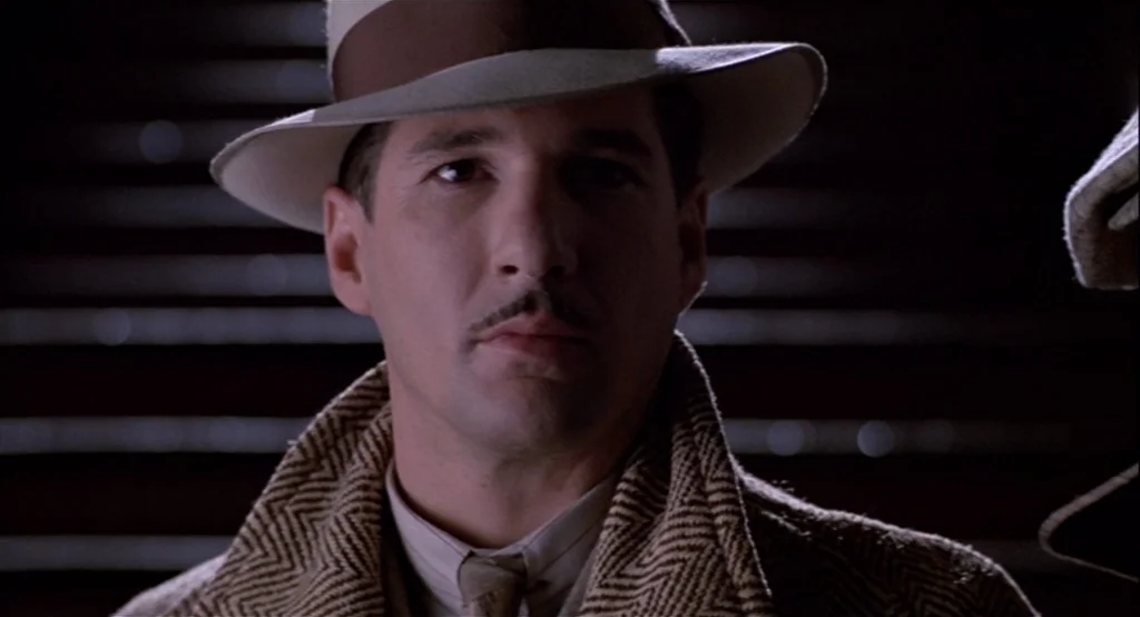 The Cotton Club (1984) movie still: closeup — A dimly lit room features horizontal blinds in the dark background. A man in a fedora and tweed…