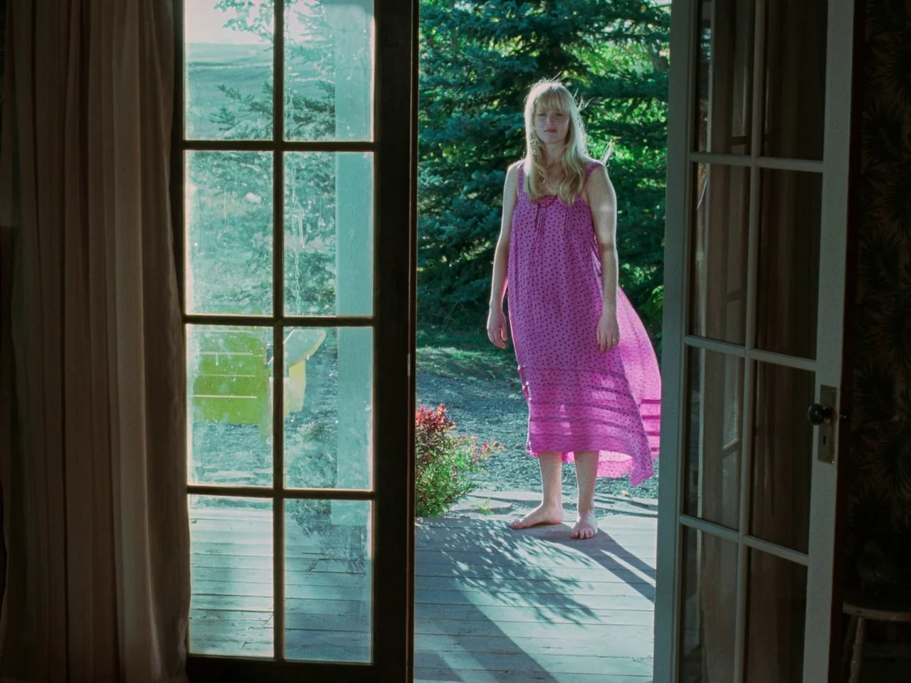 Die My Love (2025) movie still: fullbody — A woman stands on a wooden porch outside a house with glass doors. She wears a pink dress and faces the…