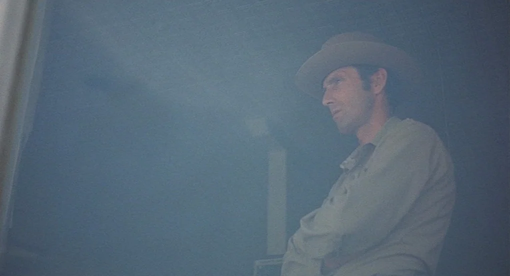 Vanishing Point (1971) movie still: medium — A dimly lit indoor space appears hazy, obscured by a translucent veil or filter. A man in a felt hat…