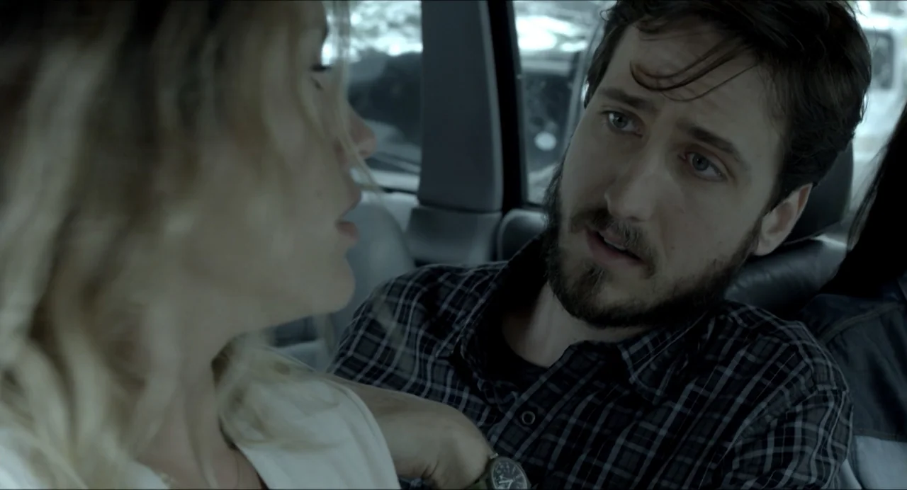 Suspension Points (2022) movie still: medium — The interior of a car during the day with natural light from the window. A bearded man in a plaid…