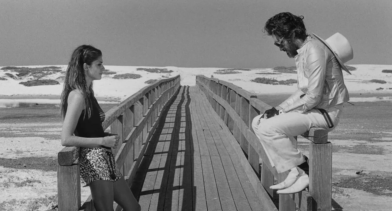 The Legend Of Kaspar Hauser (2012) movie still: fullbody — A wooden footbridge stretches across a pale, sandy landscape with sparse vegetation. A…