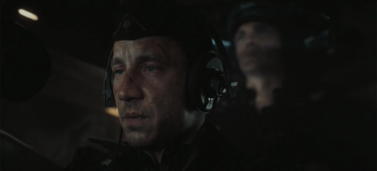 The Tank (2025) movie still: closeup — The cramped interior of a military vehicle features dark metal walls and minimal lighting. A man in a uniform…