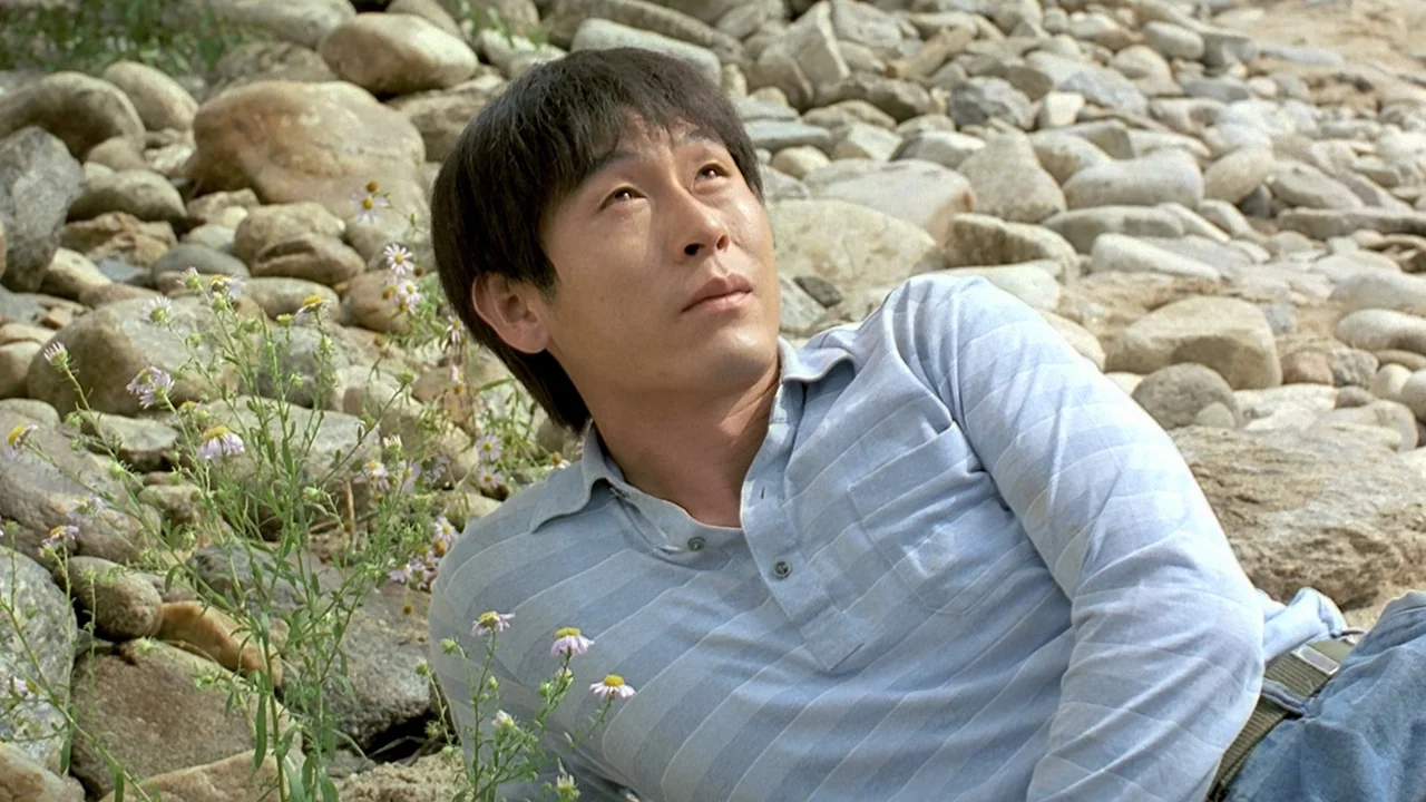 Peppermint Candy (1999) movie still: medium — A rocky riverbank is filled with rounded stones and small patches of wild flowers. An East Asian man…