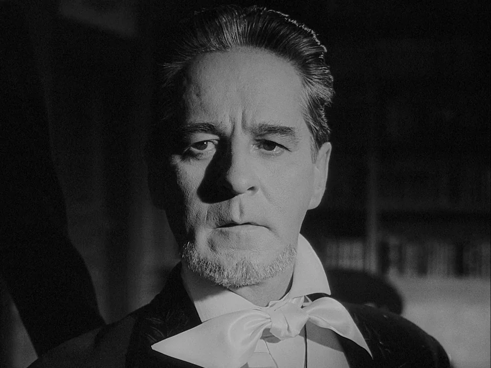 The Magician (1958) movie still: closeup — A dimly lit library interior with blurred shelves visible in the dark background. A middle-aged man in…